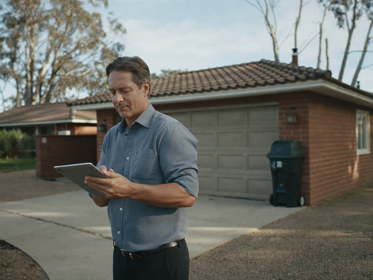 A valuer standing outside a suburban brick house, making notes on a tablet.
